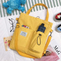 Yellow Harajuku canvas tote bag with multiple pockets and shoulder strap on bed with glasses and toys