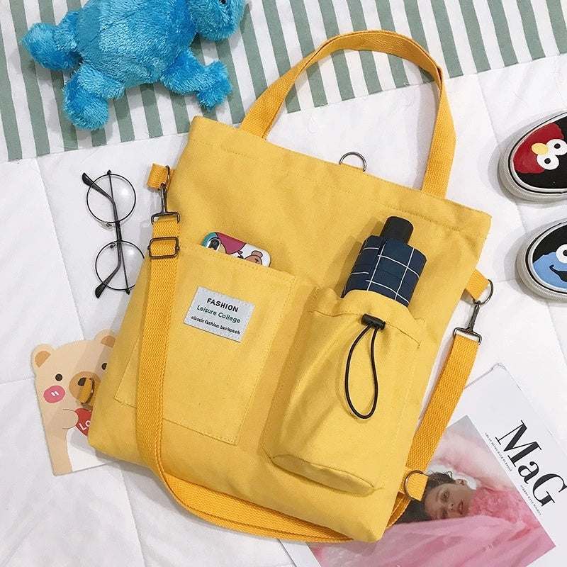 Yellow Harajuku canvas tote bag with multiple pockets and shoulder strap on bed with glasses and toys